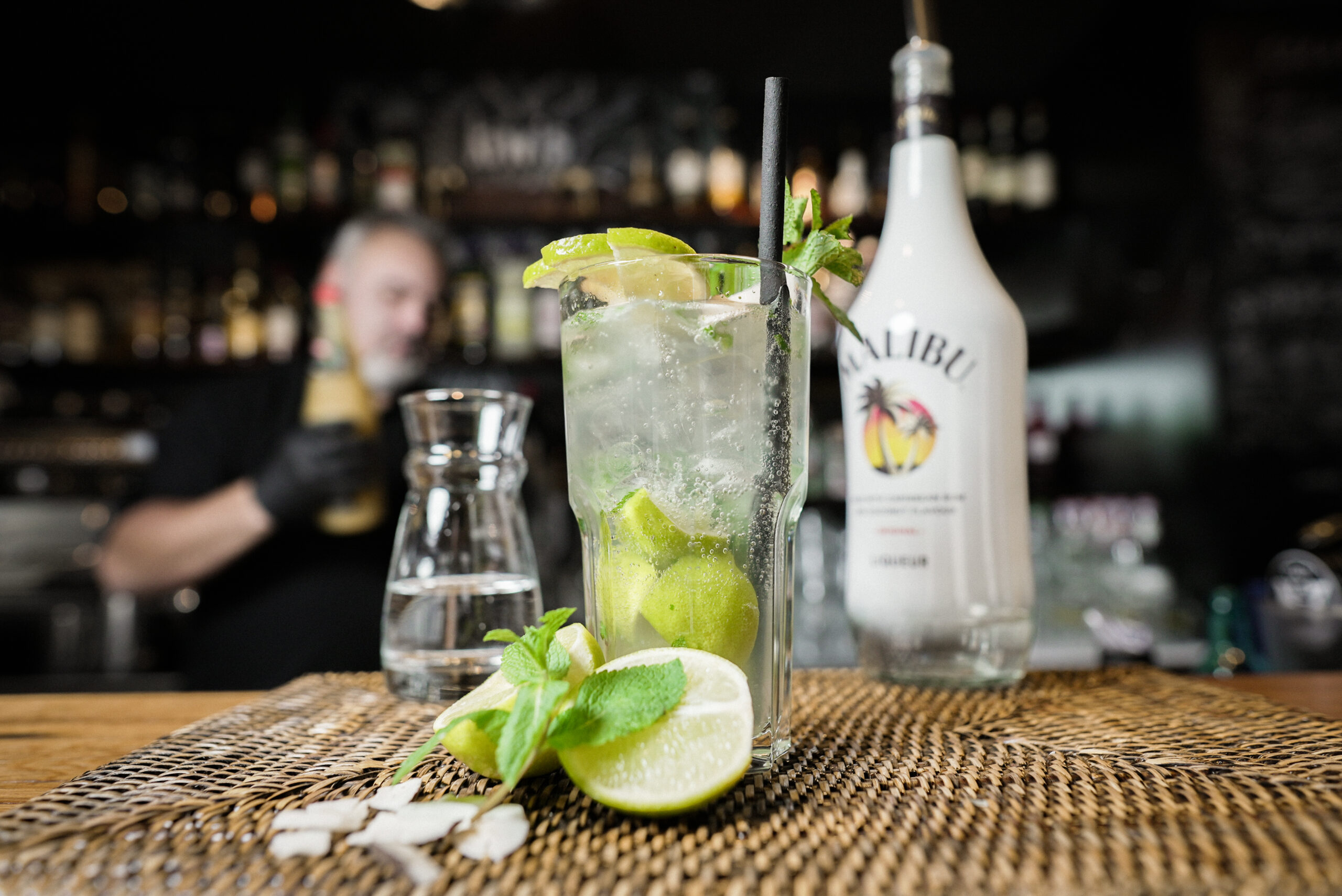 Cocktail angerichtet im großen Glas mit Limetten und Mize, im Hintergrund eine Bar mit Malibu und Barkeeper