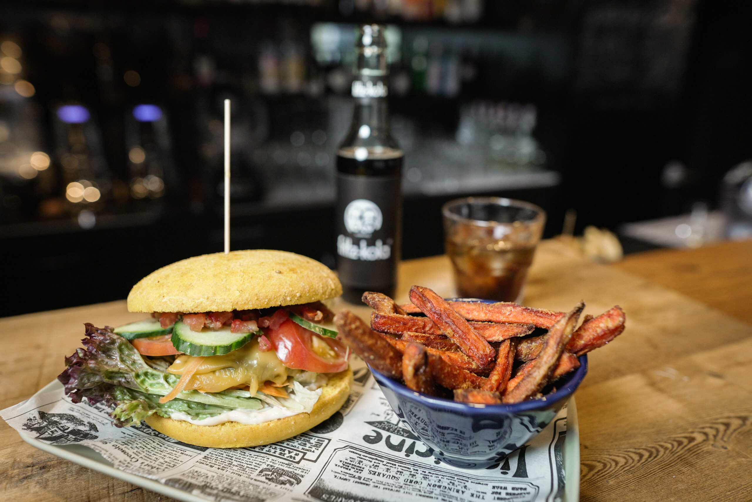 Burger mit Gemüse, Käse, Fleisch und Yellow Burger Bun an einer Bar, daneben Süßkartoffel Fries und im Hintergrund fritz-kola